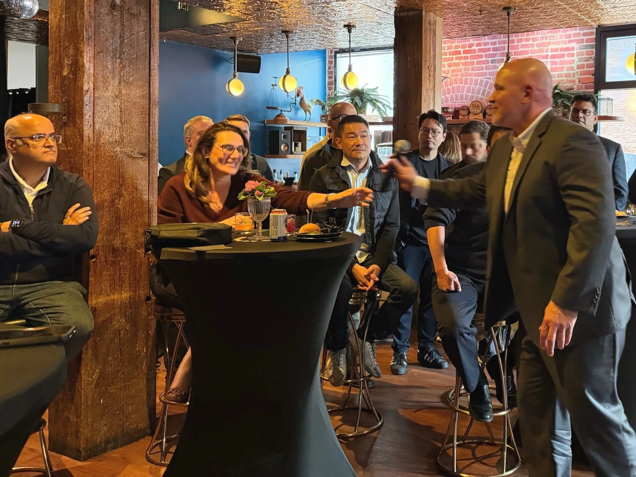 A speaker with a microphone addresses an audience clustered around tall cocktail tables at the SaaSpocalypse event